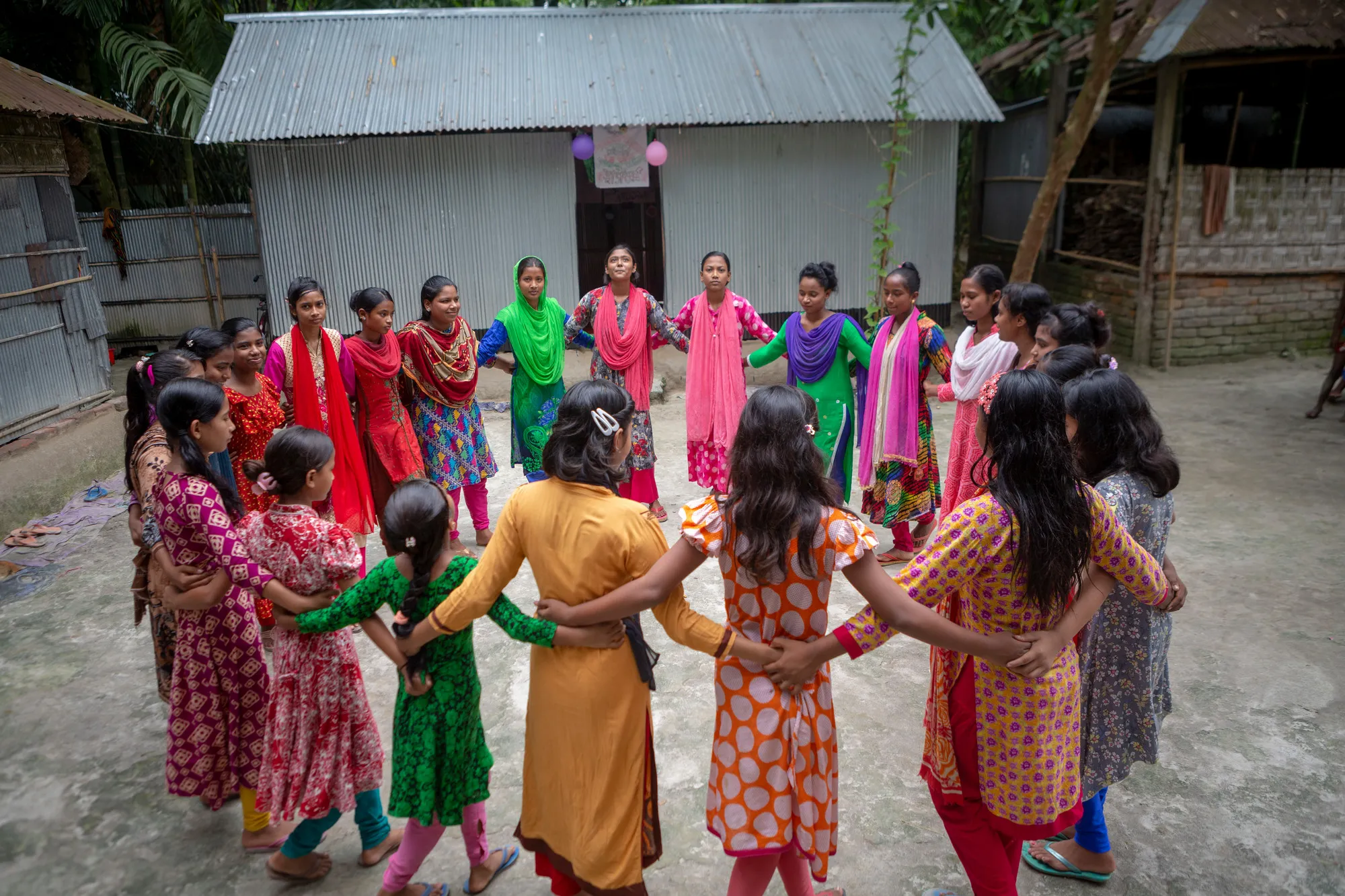 A group of adolescent girls stand in a circle outdoors in a village.