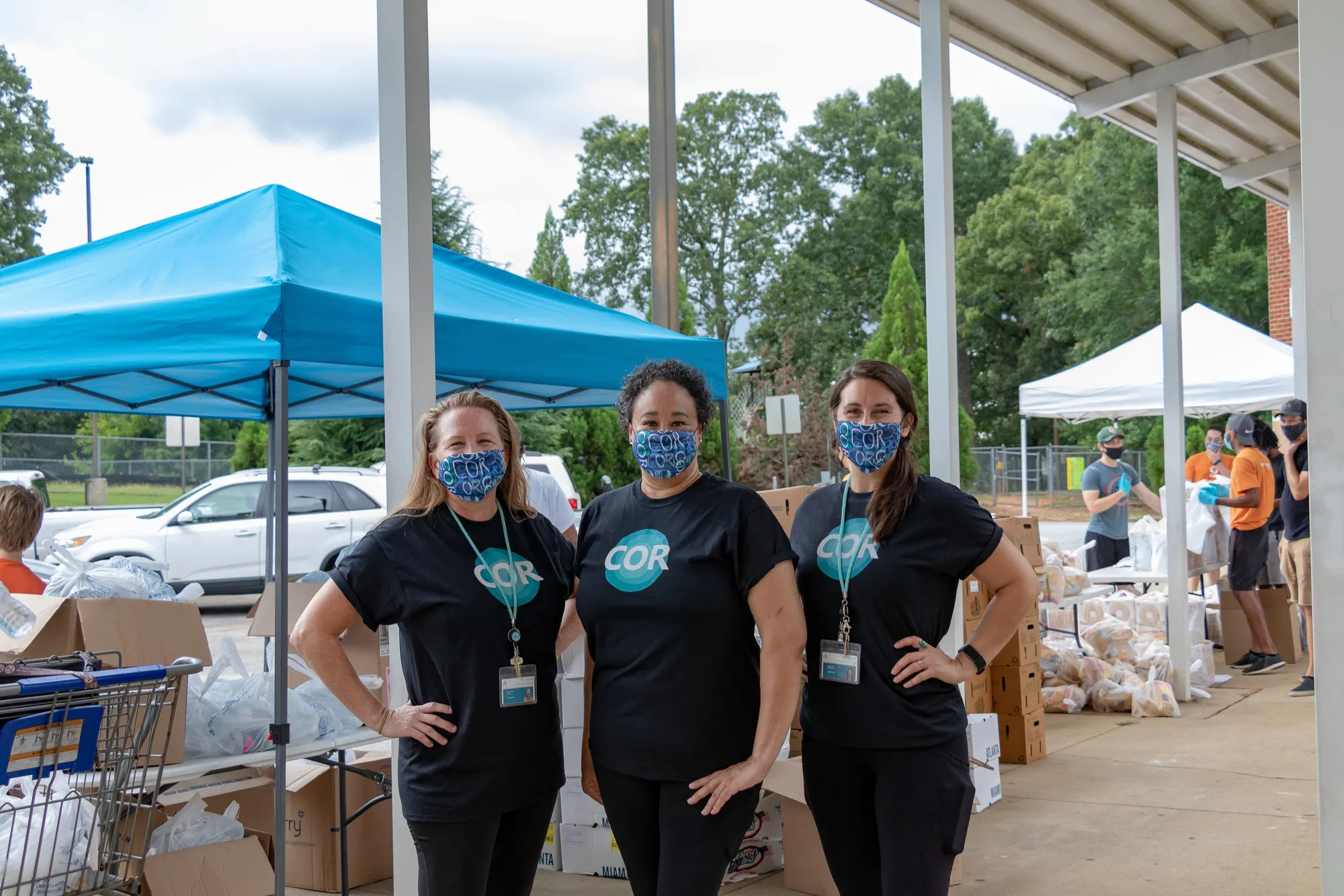 Three women in face masks stand behind a tent set up at a food distribution site.