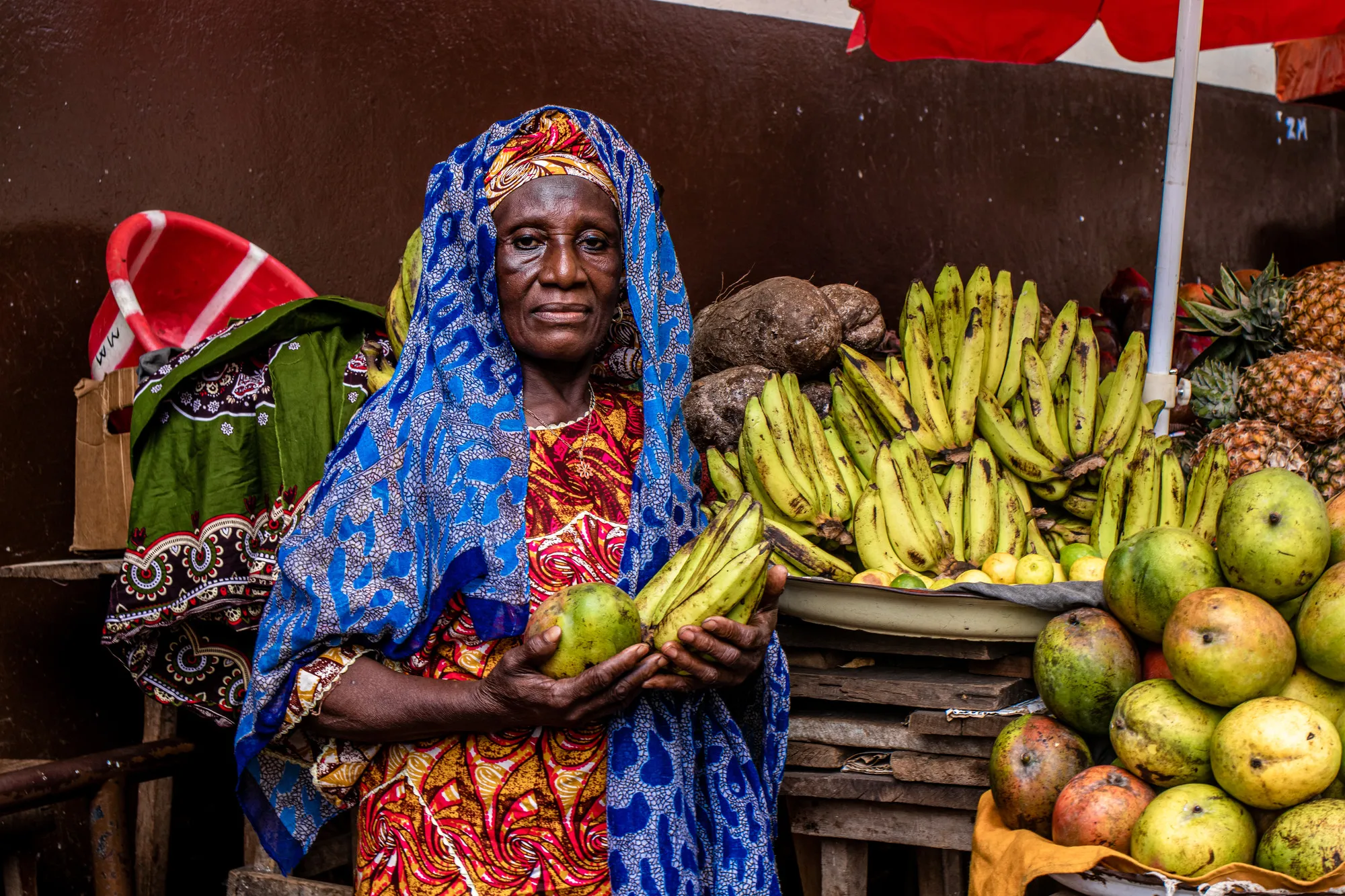 A woman at a fruit stand holds bananas and other fruits.
