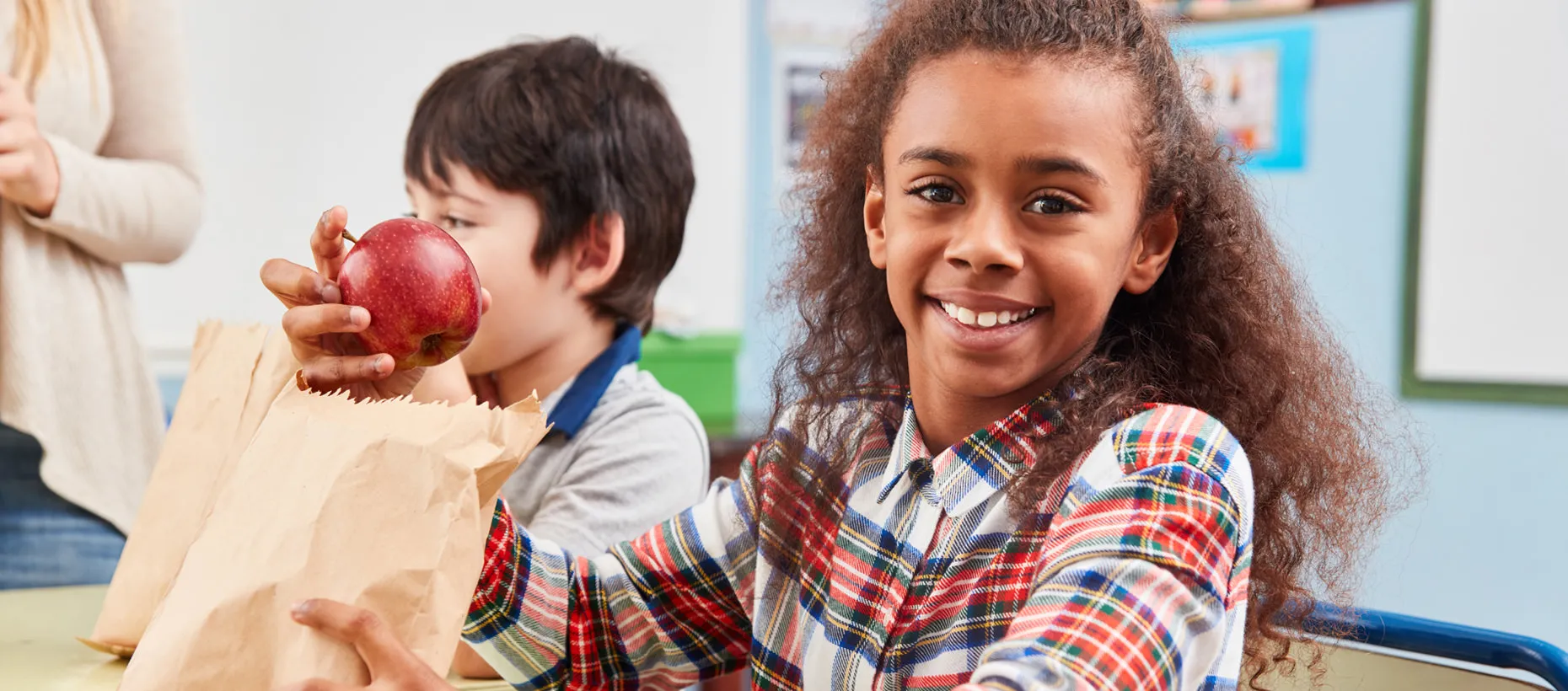 A girl smiles while setting at a school desk while holding an apple.