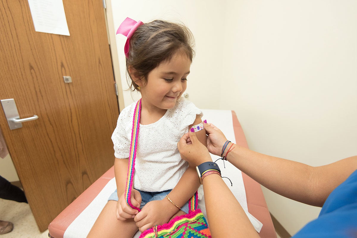 A young girl with her hair pulled back in a pink ribbon smiles while someone places a cartoon band-aid on her arm.