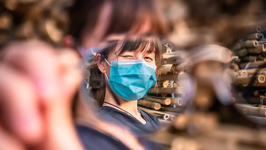 Uma mulher usando uma máscara facial médica azul pisca para a câmera. Seu rosto está em foco nítido, mas tudo ao seu redor está borrado.