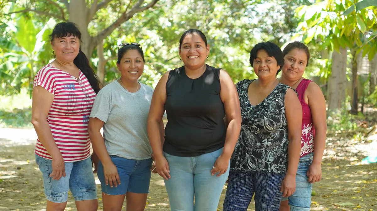 A group of Peruvian women stand together and smile outside under a large tree.