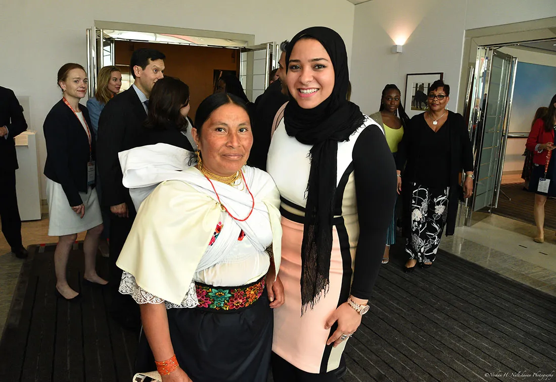 A young woman wearing a black head scarf stands next to a woman wearing a white top and a black shirt with floral embroidery.