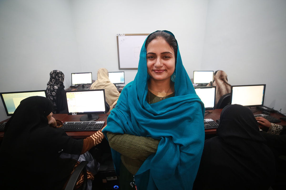 Una mujer que lleva un pañuelo azul se encuentra frente a un grupo de mujeres que trabajan con computadoras.