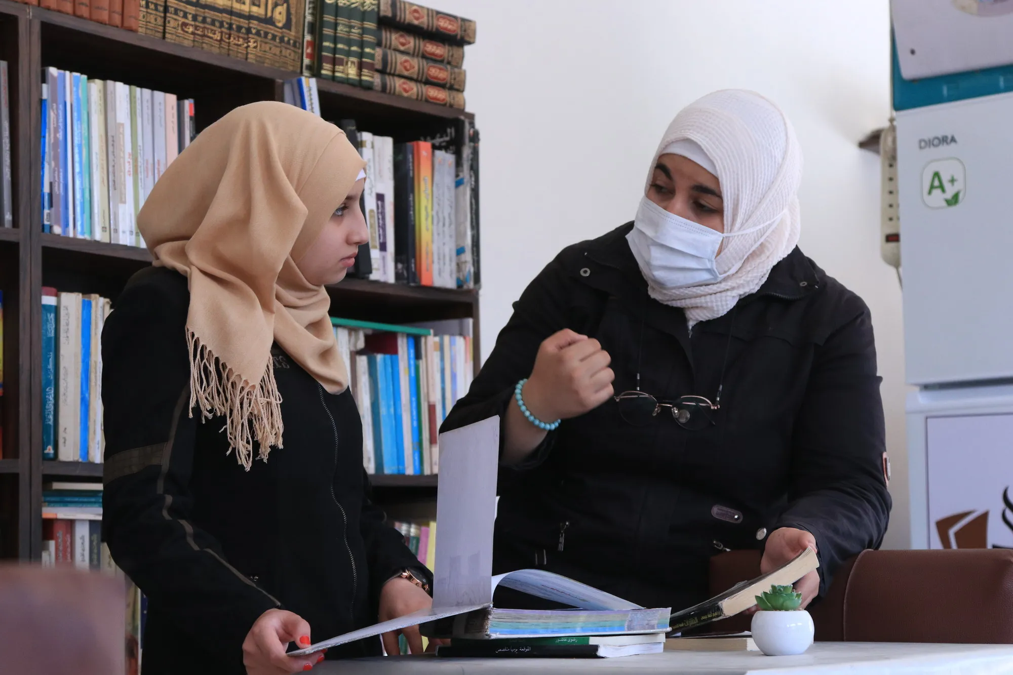 A woman in a face mask speaks with a girl in a library.