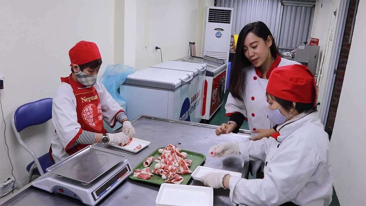 A Vietnamese woman wearing a white and red chef's apron speaks to two food workers, both wearing red hats and blue masks, while they prepare meat.