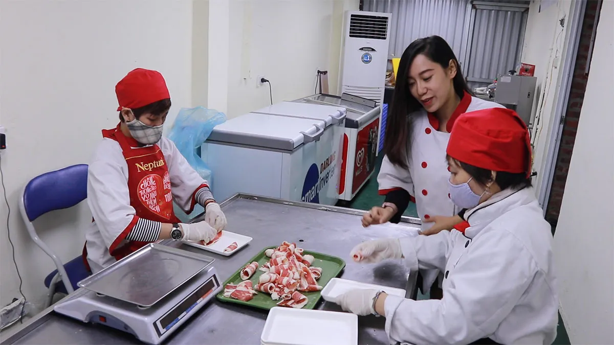Una mujer vietnamita con un delantal de chef blanco y rojo habla con dos trabajadores de alimentos, ambos con sombreros rojos y máscaras azules, mientras preparan carne.