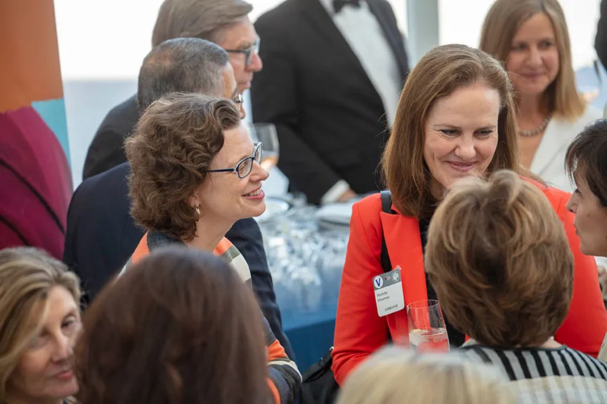 Michele Flournoy, wearing a bright red suit jacket, and Michelle Nunn, wearing an orange, gray, and white striped sweater, talk with attendees.