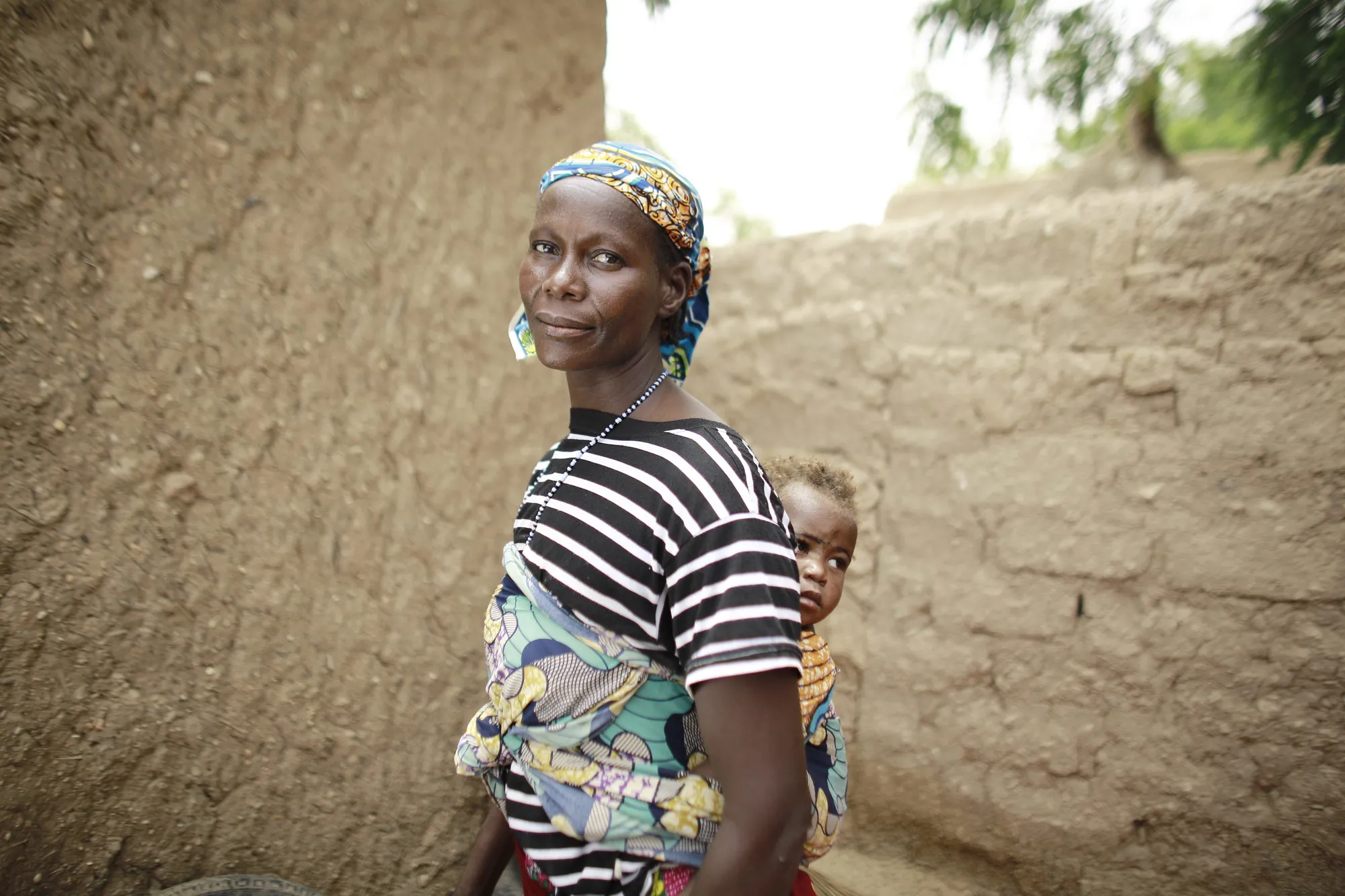A woman with a child on her back stands outside in front of a wall