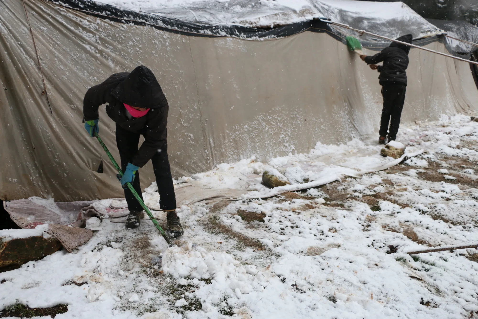 Two children shovel snow outside a tent in a camp.