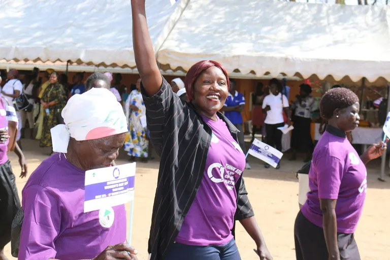 A woman wearing a purple shirt smiles and raises her right arm. She is walking with three other women, all wearing matching purple shirts.