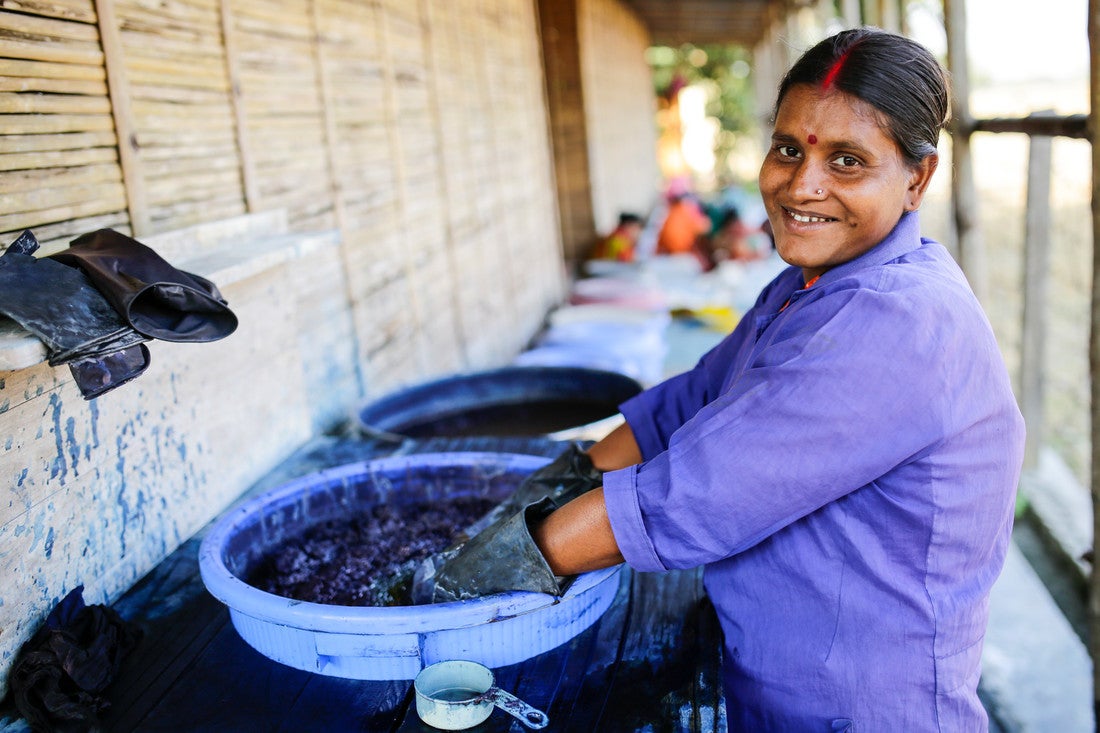 A woman smiles while plunging her gloved hands into a blue bucket filled with dark blue dye. She is smiling at the camera and wearing a blue long-sleeved shirt with the sleeves rolled up.