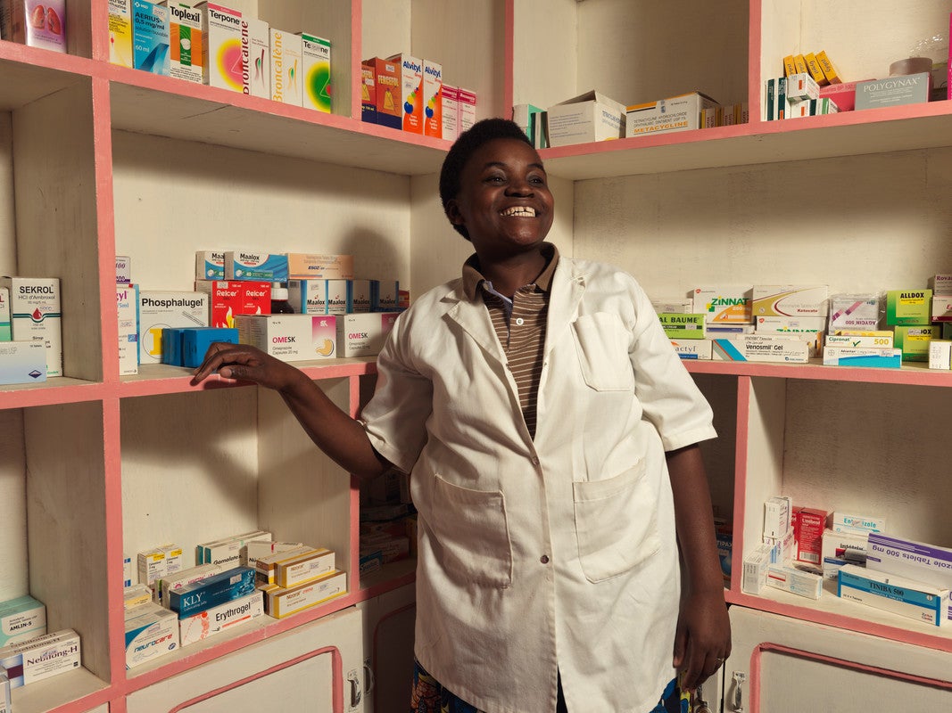 A woman wearing a white lab coat smiles while standing in a pharmacists office. She is leaning against white shelves stacked with medicine.