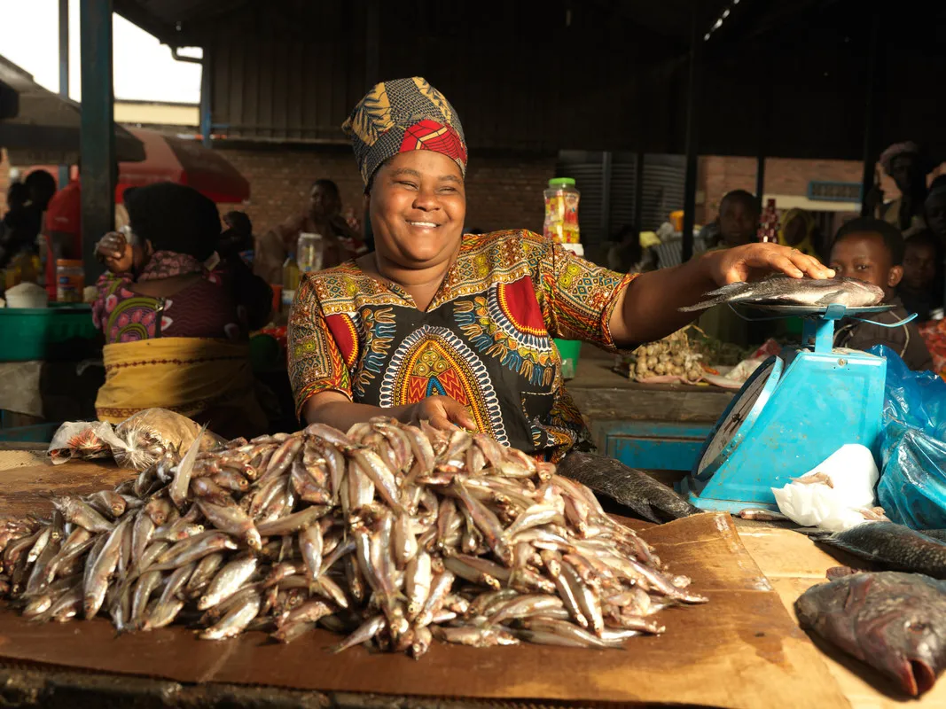 Una mujer vestida con una camisa estampada y un sombrero sonríe en una mesa llena de peces. Ella sonríe y apoya la mano sobre la mesa.