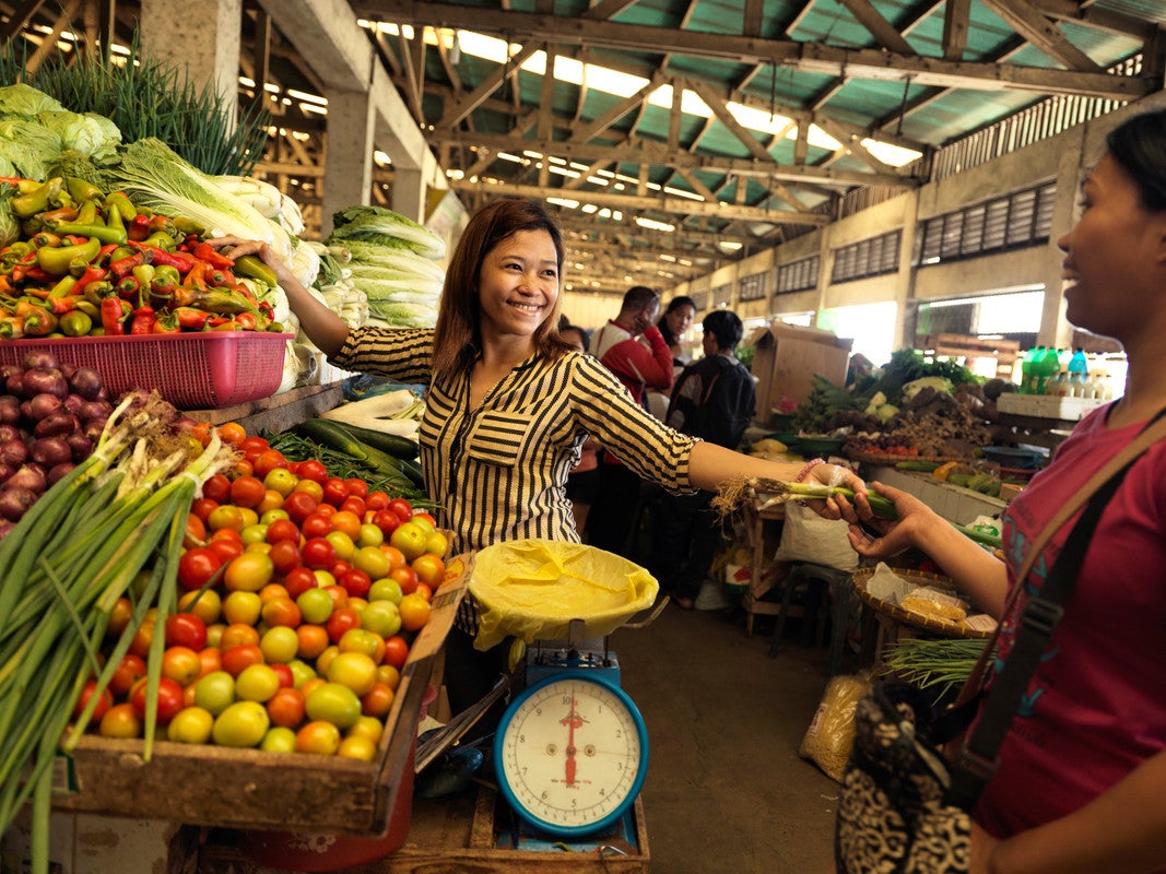 Una mujer con una camisa a rayas sonríe mientras le entrega una verdura a alguien. Ella está en un mercado de frutas junto a grandes barriles de productos.