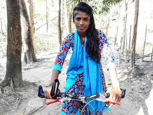 A lady wearing a colorful a dress and a blue scarf is riding a bike.