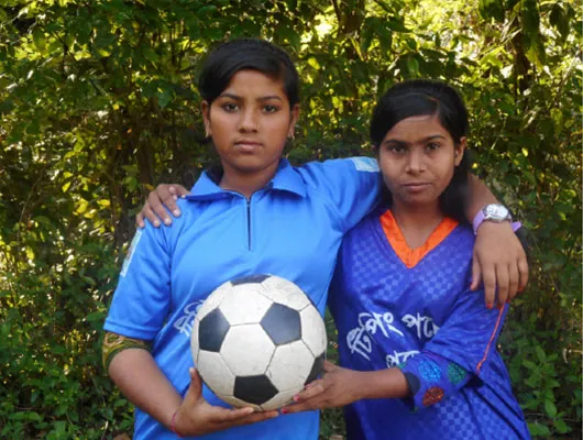Two girls wearing royal blue soccer jerseys hold a soccer ball together.