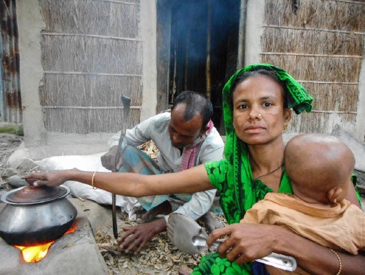 A woman and man sit outside cooking. The woman is holding a baby and looking at the camera.