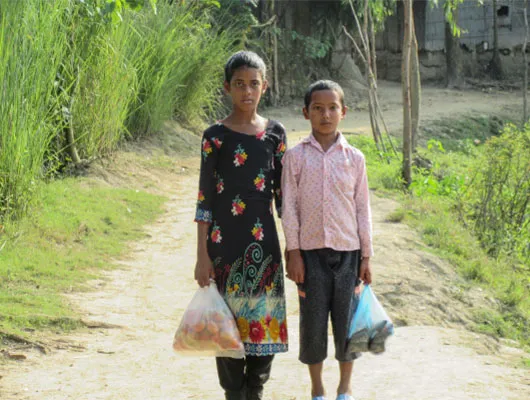 Two young children stand next to each other on a dirt path. They are both carrying bags of groceries.