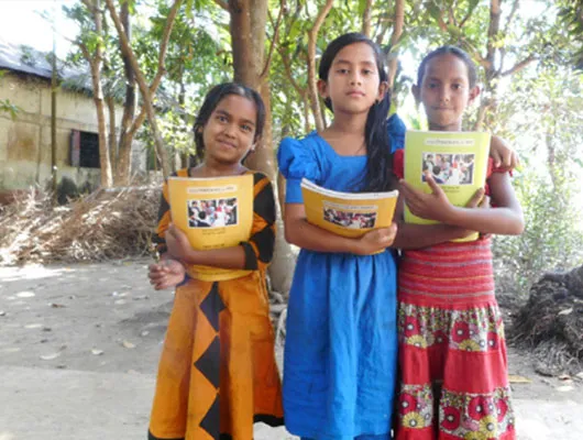 Three young girls wearing boldly colored dresses stand together while holding yellow schoolbooks.