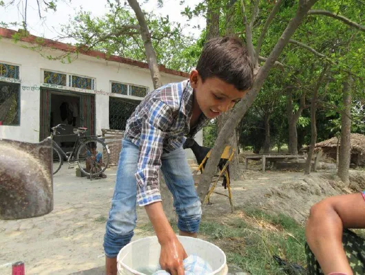 A young boy wearing jeans in holding a piece of cloth from a bucket.