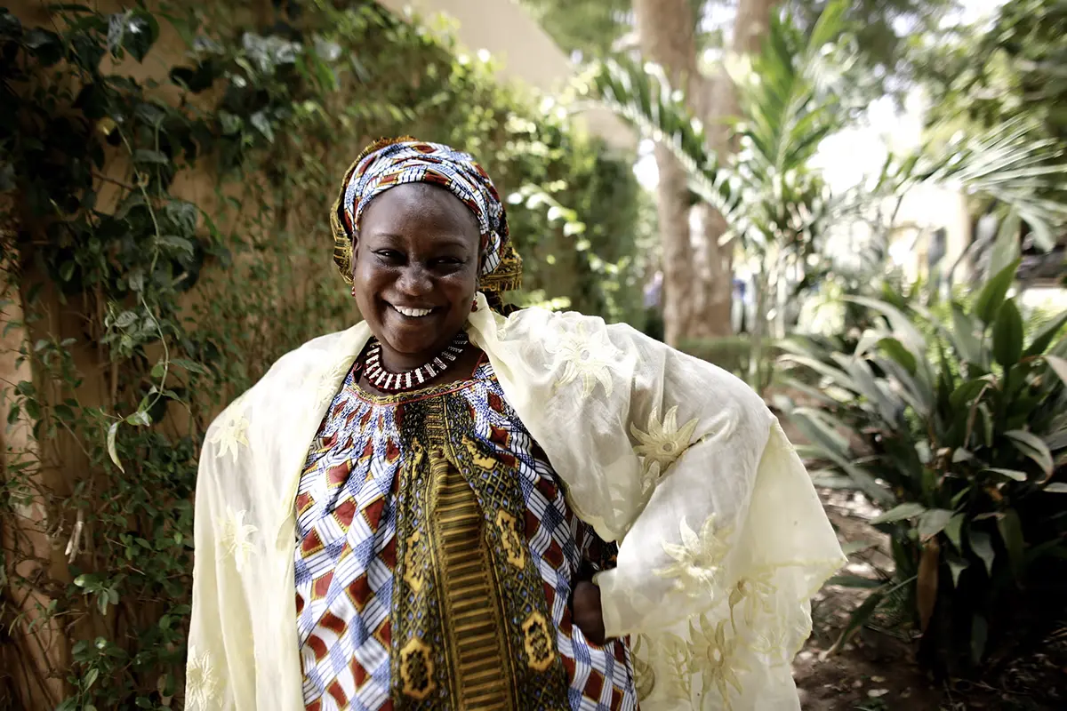 A woman wearing a patterned shirt and headscarf smiles with her hands on her hips.