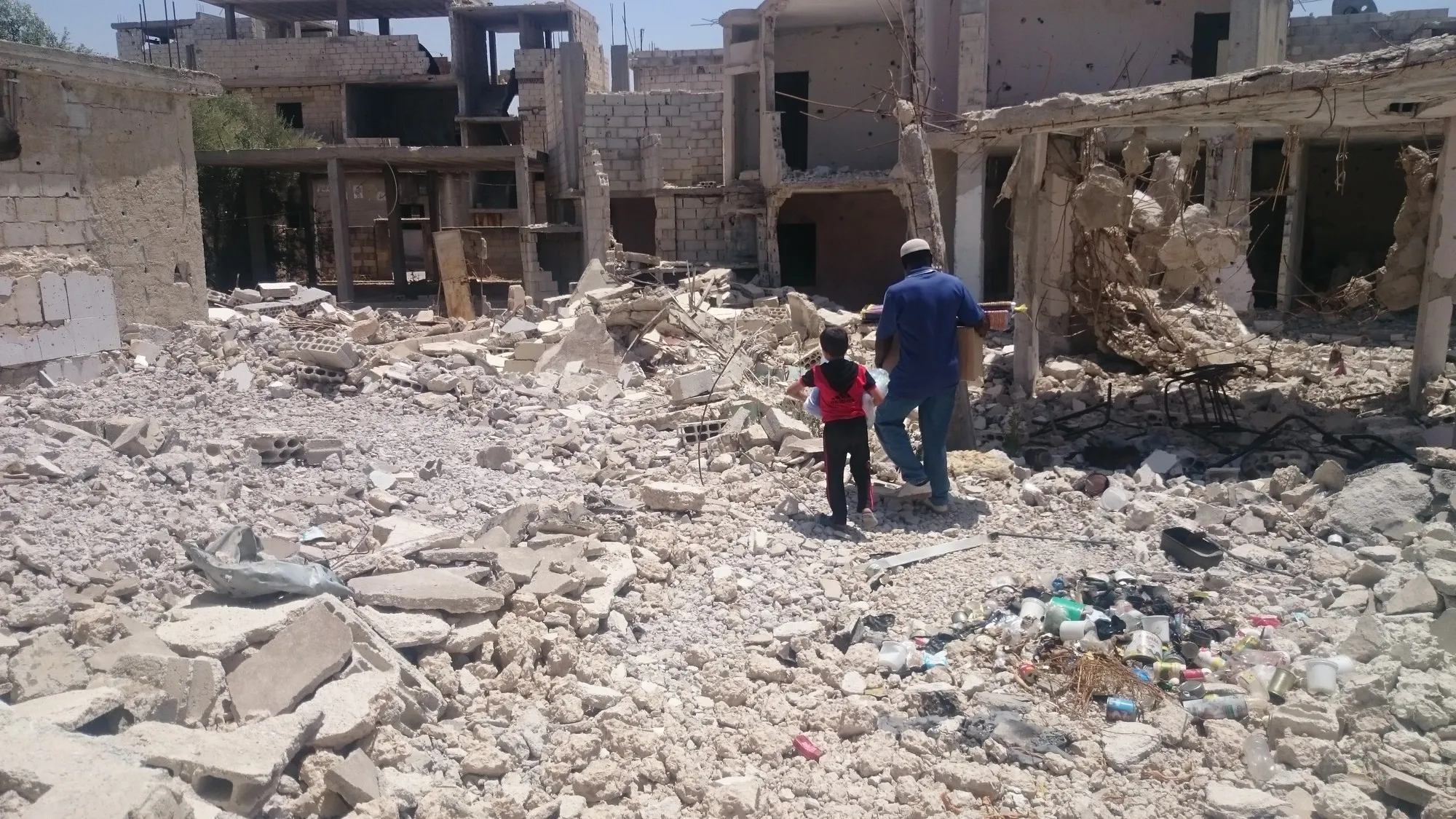 A man and a child walk through the rubble of a destroyed building.