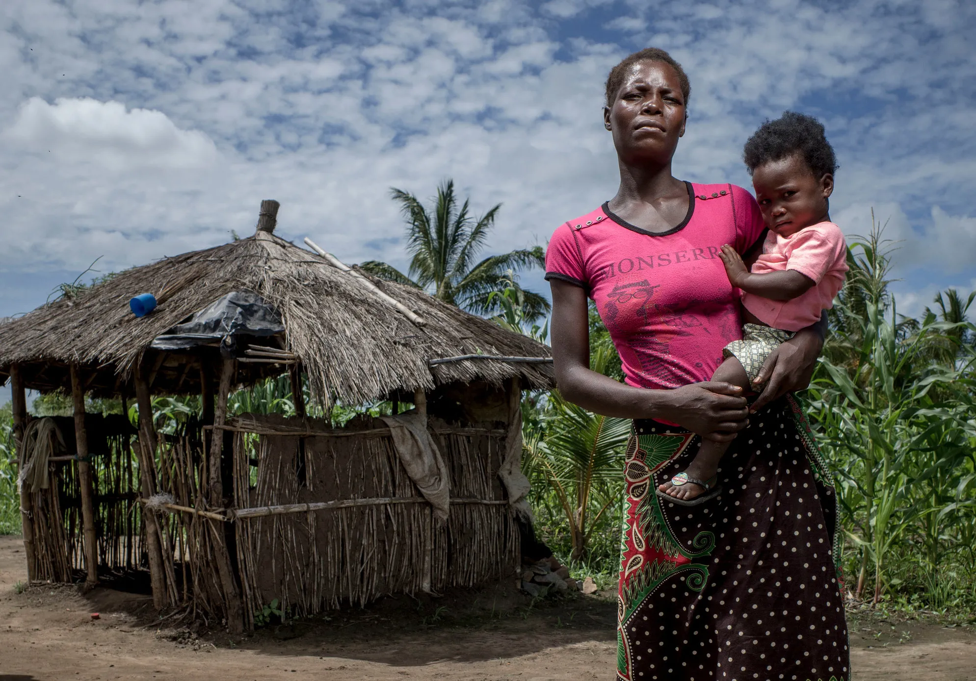 A woman holds her daughter in front of a wooden hut.