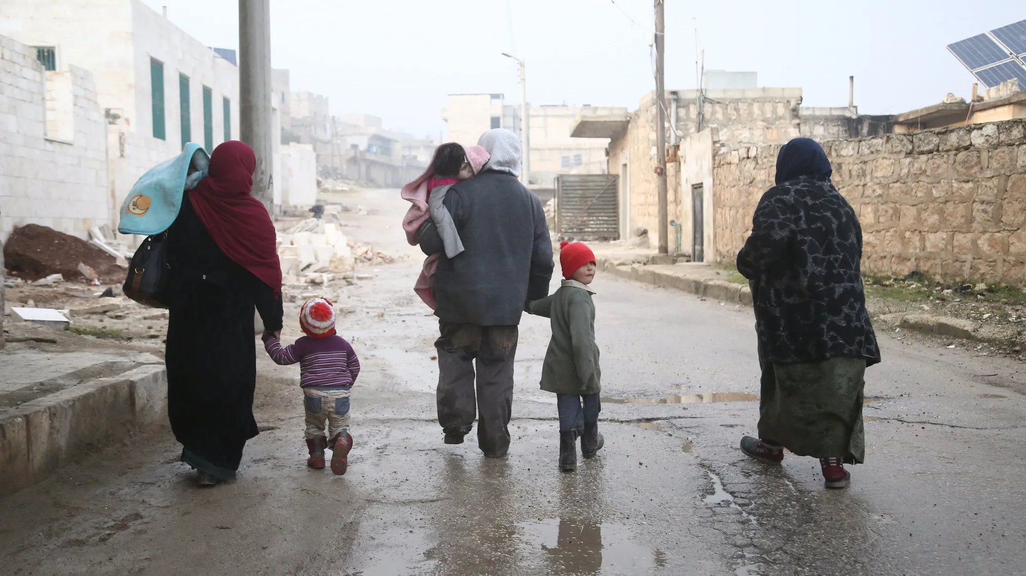 A group of children and adults walk in winter clothes in a bombed out city.
