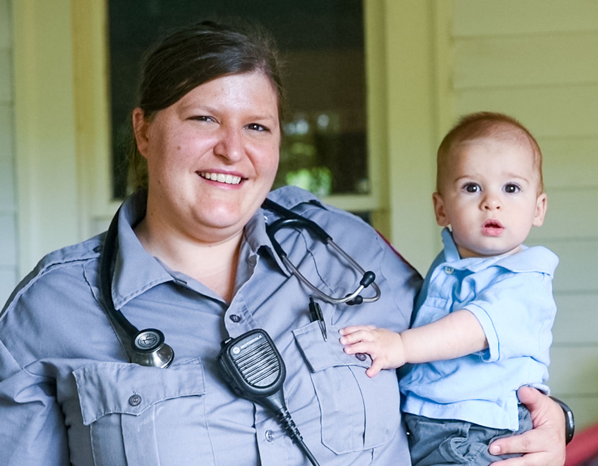 A woman emergency healthcare worker smiles in uniform while holding her young child.