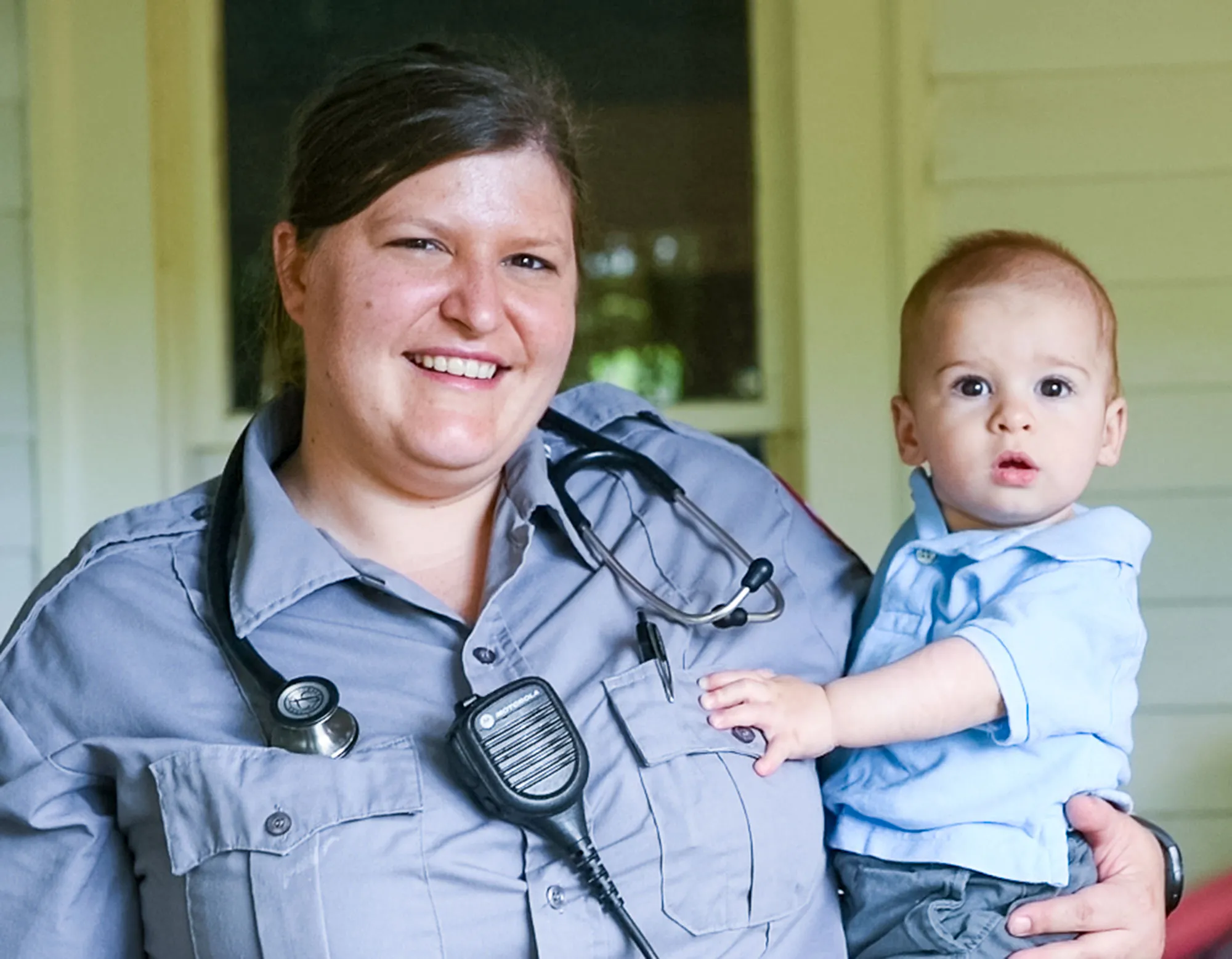 Una trabajadora de la salud de emergencia de la mujer sonríe en uniforme mientras sostiene a su niño pequeño.