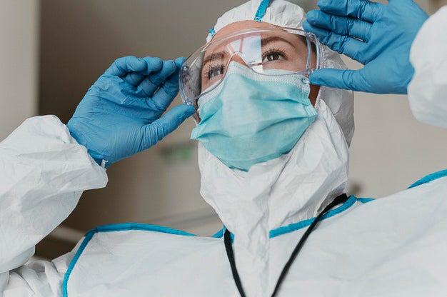 A woman healthcare worker adjusts protective glasses on her face. She is wearing a white protective suit, a blue facemask, blue gloves, and clear protective goggles.