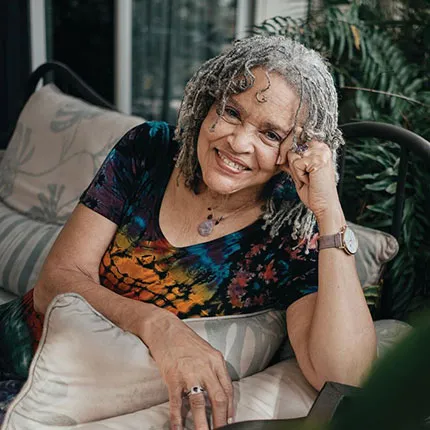 Charlayne Hunter-Gault smiles while sitting on a couch and resting her arms on the sidearm.