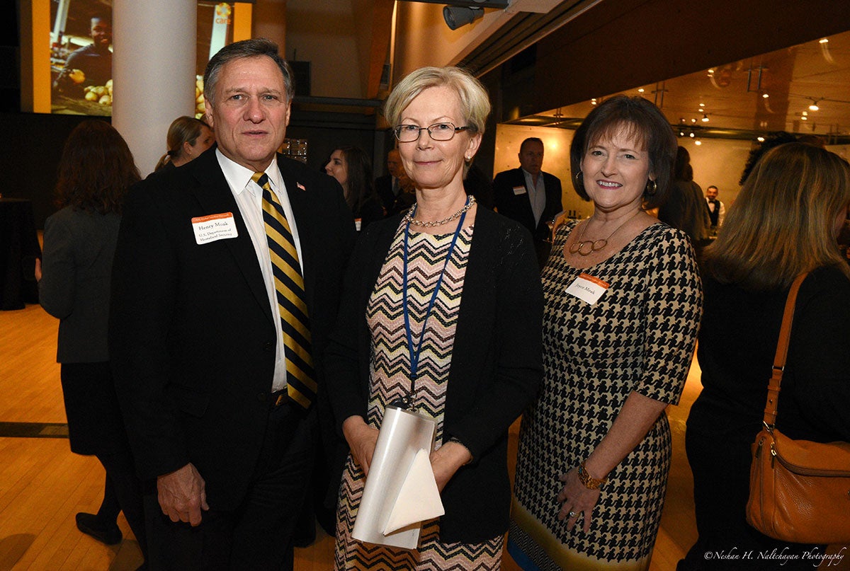 Ambassador Kristi Kauppi stands in the middle of a man in a black suit and a woman in a yellow and black dress