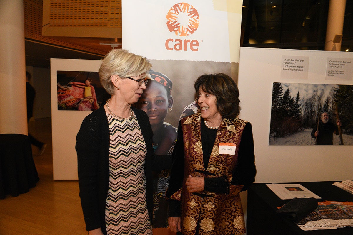 Ambassador Kristi Kauppi and a woman in colorful clothing are standing and talking to each other in front of a CARE banner