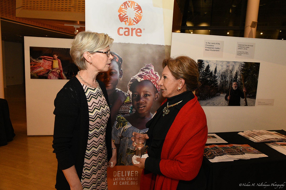 Ambassador Kristi Kauppi and a woman in a black dress and a red coat stand together in front of a CARE banner