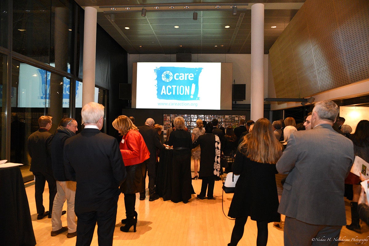 Multiple people in formal attires are standing while there is a CARE Action logo projected on the screen