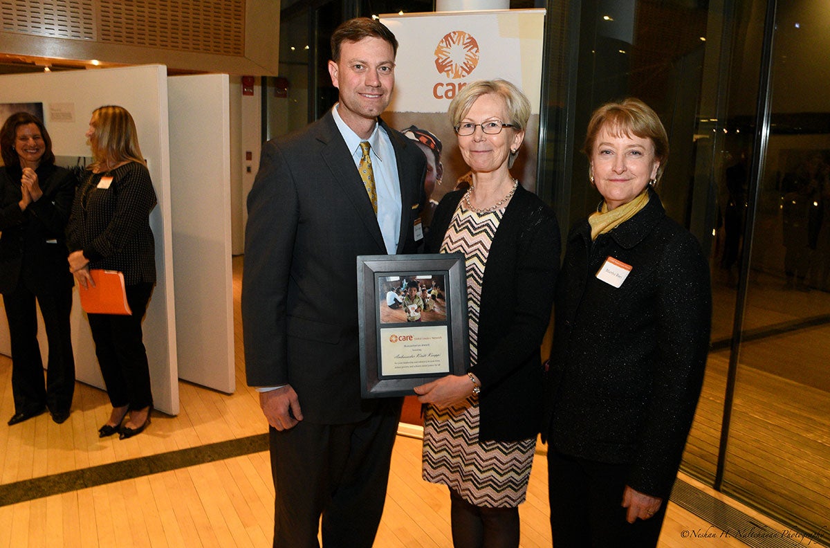 Ambassador Kristi Kauppi is holding a CARE award standing between a man in a black suit and a woman in a black jacket in front of a CARE banner