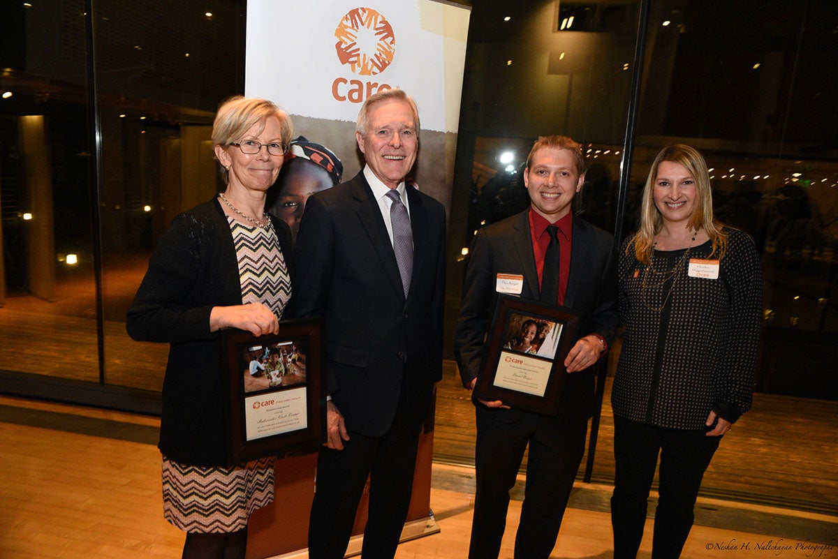 Ambassador Kristi Kauppi is holding a CARE award standing next to two men in black suits, one of them holding another CARE award, and a woman in a black and white shirt in front of a CARE banner