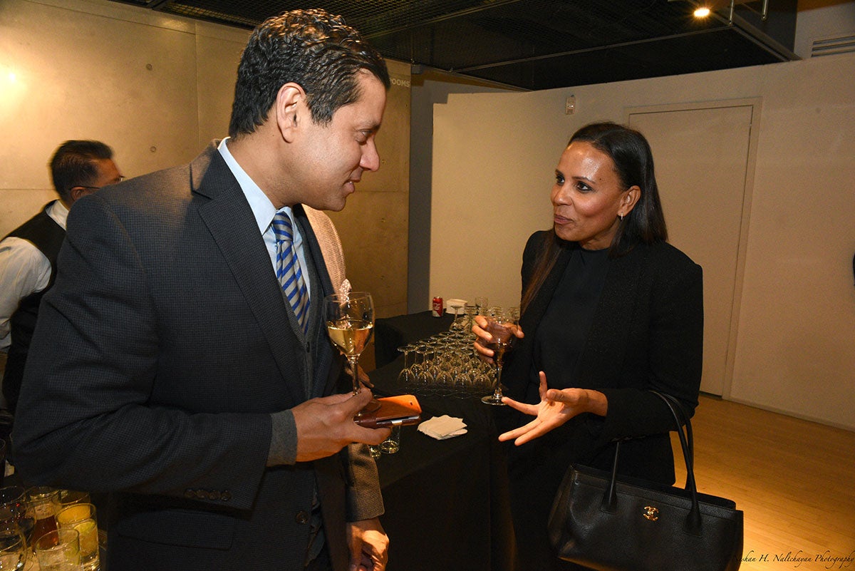 A man in a black suit is talking to a woman in a black suit. Both are holding glasses of a beverage
