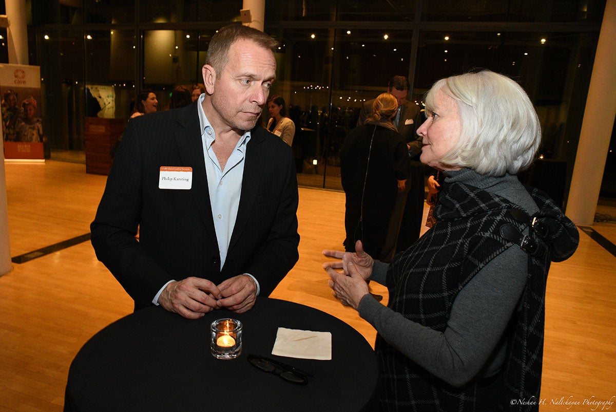 A woman in a grey shit and a black and white scarf is talking to a man in a black suit, both standing next to a table with a small candle on top of it