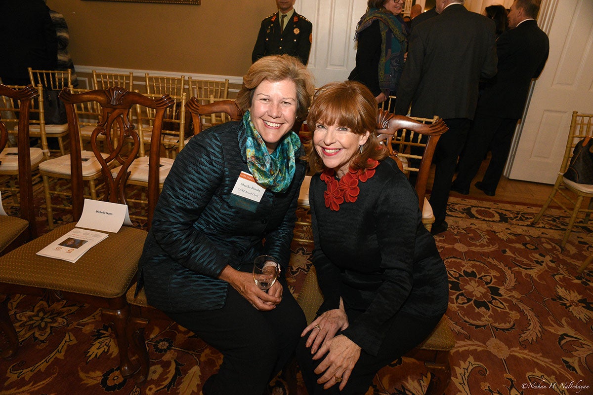 Two women, one in a dark green and another in black clothing, are smiling while sitting on chairs