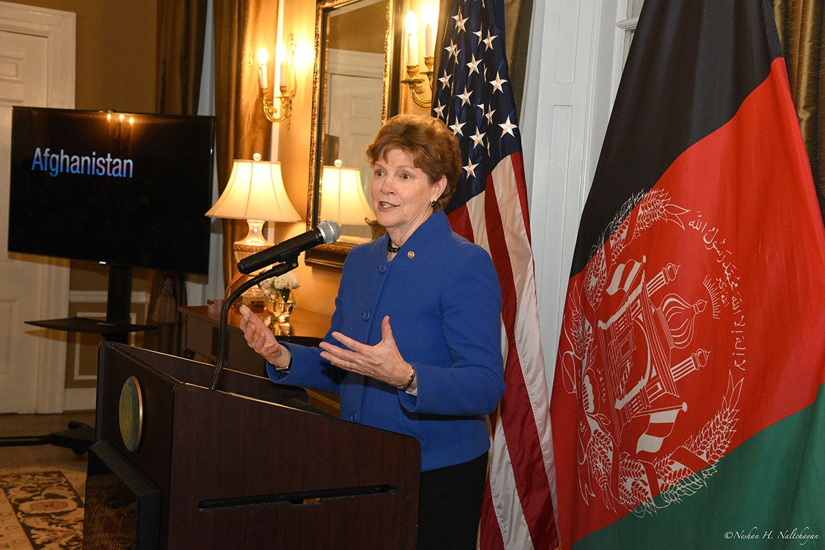 A woman in a blue jacket is holding a speech with both the American and Afghan flags in the background