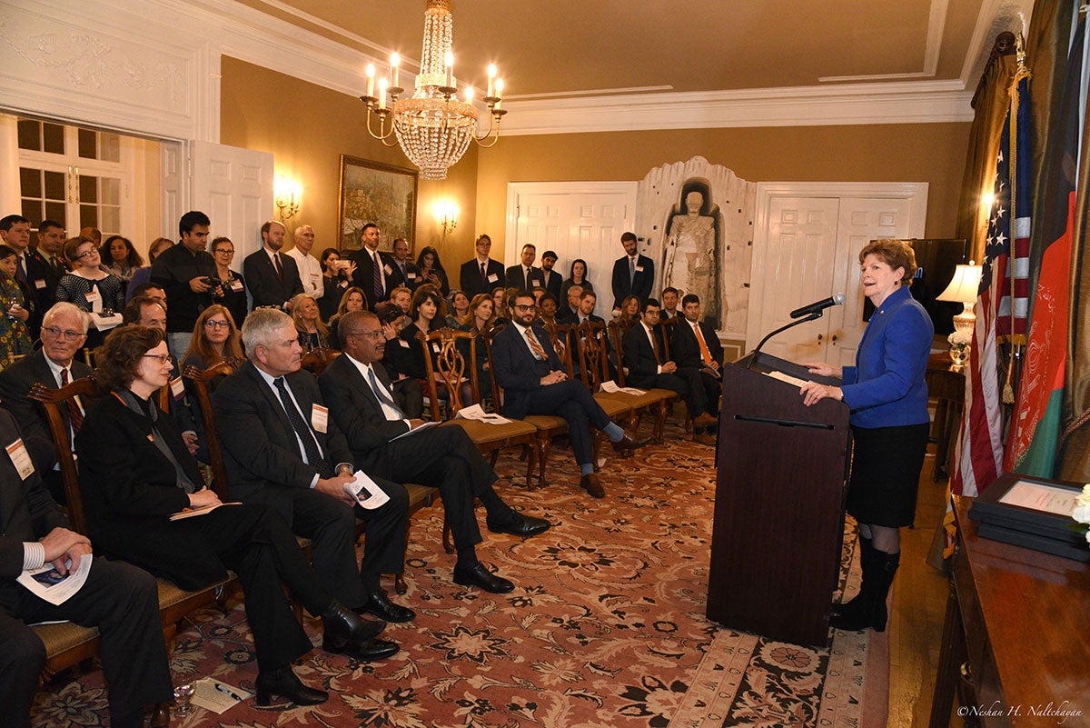 A woman in a blue jacket is holding a speech in front all guests who are sitting on chairs