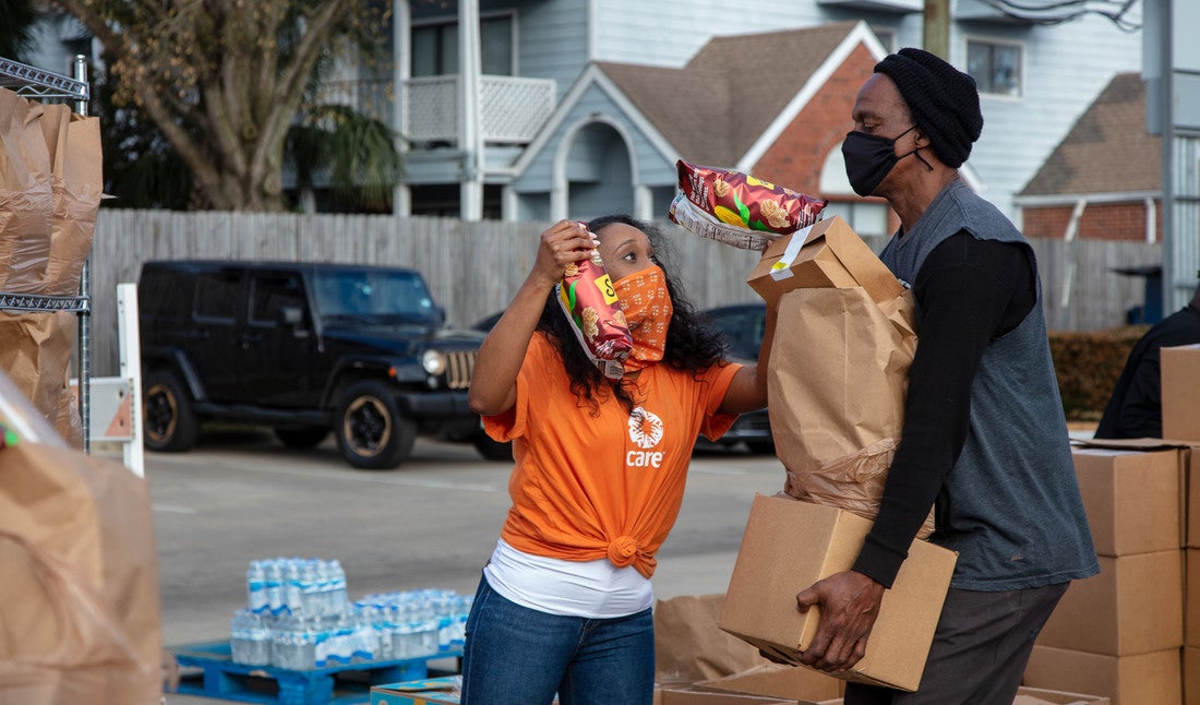 A woman wearing an orange CARE t-shirt and matching face mask holds two bags of chips up to a man carrying a cardboard box with a large paper bag stacked on top. Behind them are large stacks of plastic water bottles.