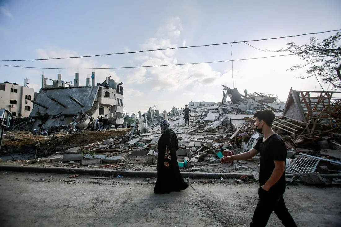 A woman wearing dark clothing walks by the ruins of a destroyed building.