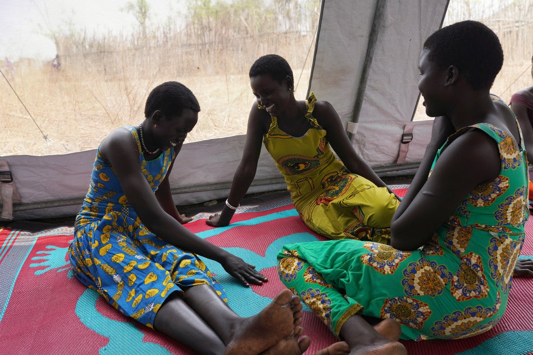 Three adolescent South Sudanese girls smile and talk together while seated on a rug inside a large tent.