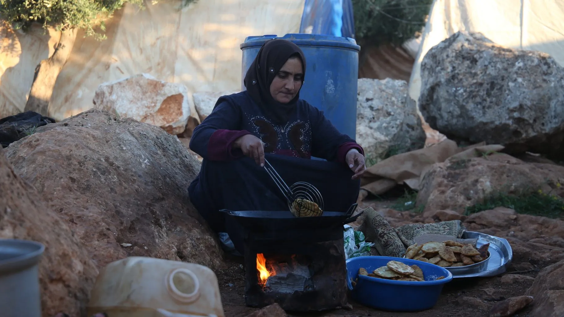 Une Syrienne vêtue de vêtements sombres cuisine au-dessus d'un feu ouvert dans un camp de déplacés