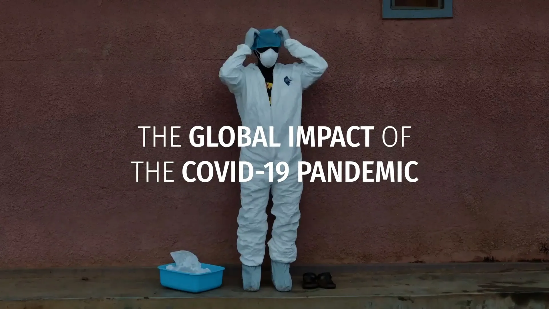 A screenshot of a video that shows a frontline health worker wearing a white suit and mask with light blue gloves and booties. Text is overlaid that says 